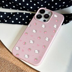 Może przedstawiać: R&oacute;żowe etui na telefon z wzorem twarzy Hello Kitty, r&oacute;żowych kokardek i truskawek. Etui ma gładką fakturę i podwyższoną krawędź chroniącą ekran i aparat telefonu. Zaprojektowane dla smartfona z trzema obiektywami.
