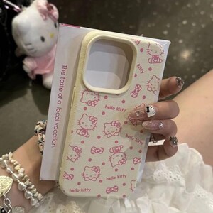 May include: A cream phone case adorned with a pink Hello Kitty pattern and bows. The case has a rectangular form with rounded edges and a camera aperture. The words "hello kitty" are also printed on the case. The packaging reads "The taste of a local vacation."