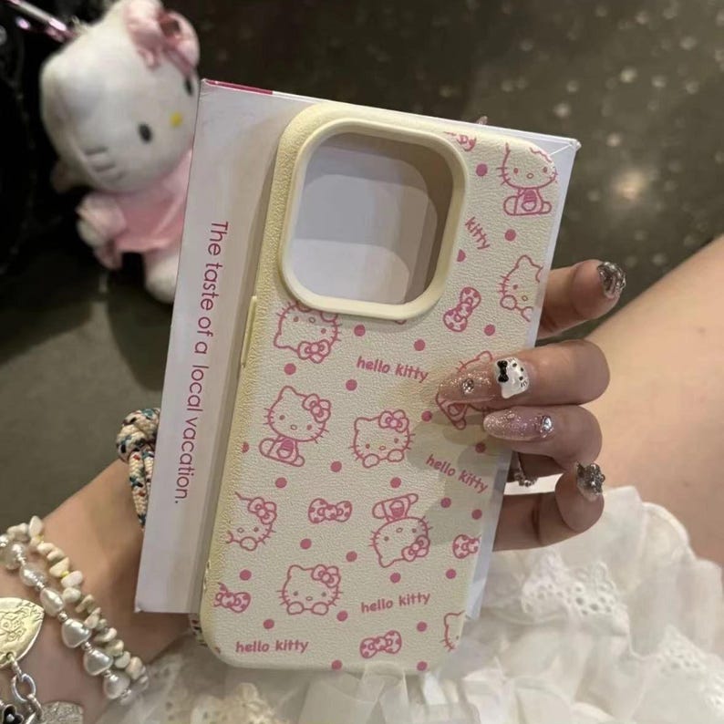 May include: A cream phone case adorned with a pink Hello Kitty pattern and bows. The case has a rectangular form with rounded edges and a camera aperture. The words "hello kitty" are also printed on the case. The packaging reads "The taste of a local vacation."
