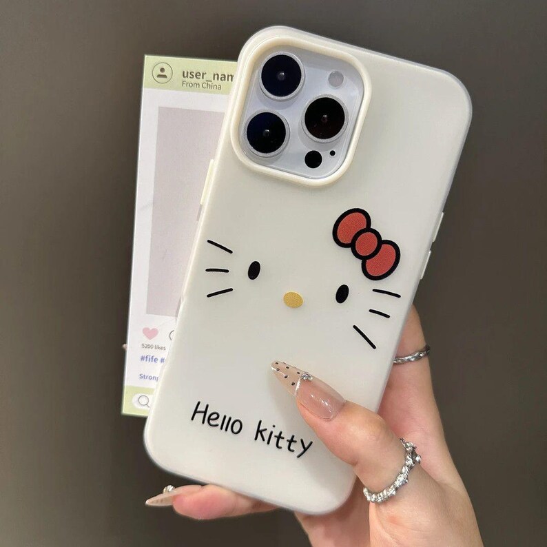 Puede incluir: Funda de tel&eacute;fono blanca con un dise&ntilde;o de Hello Kitty. La funda tiene un lazo rojo, bigotes negros y las palabras "Hello Kitty" en negro. La funda del tel&eacute;fono est&aacute; sujeta por una mano con u&ntilde;as largas y decoradas.