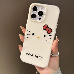 Puede incluir: Funda de tel&eacute;fono blanca con dise&ntilde;o de Hello Kitty. Presenta un lazo rojo, bigotes negros y el texto "Hello Kitty". La funda tiene un recorte para tres lentes.