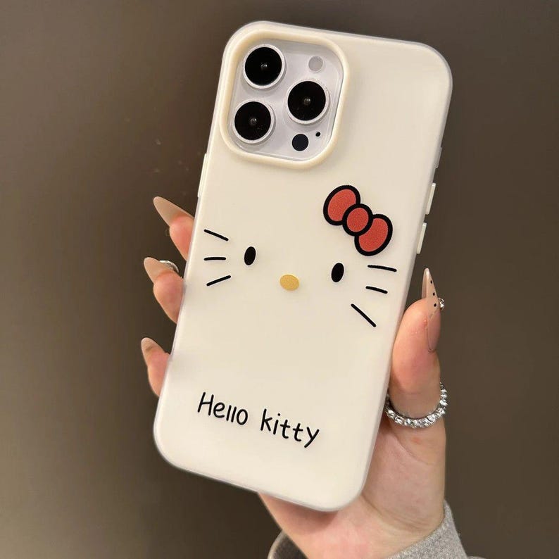 Puede incluir: Funda de tel&eacute;fono blanca con dise&ntilde;o de Hello Kitty. Presenta un lazo rojo, bigotes negros y el texto "Hello Kitty". La funda tiene un recorte para tres lentes.