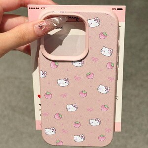 Może przedstawiać: R&oacute;żowe etui na telefon z powtarzającym się wzorem twarzy Hello Kitty, r&oacute;żowych kokardek i truskawek. Etui ma gładką fakturę i podniesioną krawędź wok&oacute;ł aparatu. Etui jest trzymane przed r&oacute;żową i białą kartą.