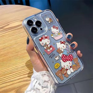 Peut inclure: Coque de t&eacute;l&eacute;phone transparente orn&eacute;e de motifs Hello Kitty et ours en peluche. L'&eacute;tui pr&eacute;sente un fond bleu clair avec des illustrations de Hello Kitty avec des n&oelig;uds, des ours en peluche et le texte "Hello Kitty" et "Sweet Day".