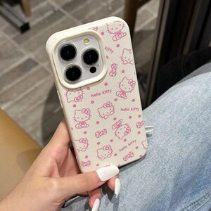 May include: A cream-coloured phone case adorned with a pink Hello Kitty design and the words "hello kitty". The case has a camera bumper and is held in a person's hand.