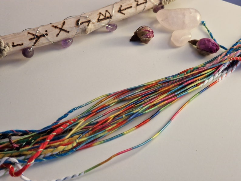 Pu&ograve; includere: Una collezione di rocchetti di filo da ricamo colorati, un bastone di legno con rune intagliate, un bocciolo di rosa rosa e un cristallo rosa.