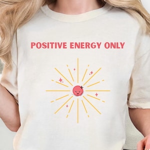 Puede incluir: Una camiseta blanca con el texto "POSITIVE ENERGY ONLY" en rojo. Debajo del texto hay un sol estilizado con una cara sonriente.