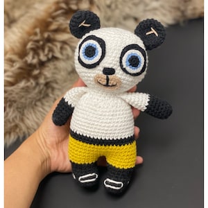 Könnte beinhalten: Häkelanleitung für ein Panda-Amigurumi, 20 cm groß, mit schwarz-weißem Fell, blauen Augen und gelber Hose. PDF-Download.