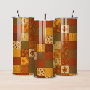 May include: Three stainless steel tumblers with lids and straws. The tumblers feature a patchwork design in shades of brown, orange, and yellow, with leaf and acorn motifs. The design evokes a fall or autumn theme.