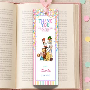 May include: A rectangular paper bookmark with a pink ribbon at the top, featuring colorful vertical stripes and illustrations of storybook characters. The text reads THANK YOU FOR BEING A PART OF MY FIRST CHAPTER, LOVE Charlie, and 12.08.2026.