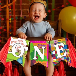 May include: A baby wearing a gold crown and a light blue long-sleeve shirt is laughing while sitting in a wooden high chair. A banner spelling "ONE" is in front of the baby, with colorful book covers forming the letters.