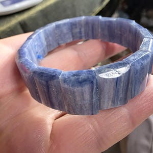 May include: A blue gemstone bracelet. The bracelet is made of rectangular, faceted blue stones. The stones have a marbled appearance with lighter blue and white streaks. The bracelet is held in a hand.