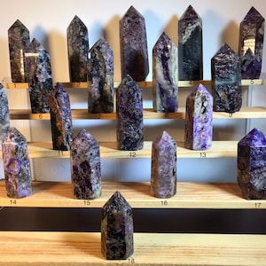 Puede incluir: Dieciocho puntas de cristal de amatista púrpura y marrón dispuestas en un estante de madera. Los cristales son de diferentes tamaños y formas, y todos están pulidos hasta obtener un acabado liso.
