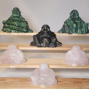 May include: Six carved stone laughing Buddha figurines on wooden shelves. The figurines are made of green aventurine, black obsidian, and rose quartz. The figurines are numbered 1 through 6.