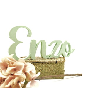 Puede incluir: Letras de madera verde deletreando el nombre "Enzo" en una cesta tejida con una pequeña rama de flores.