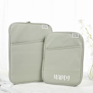 Puede incluir: Dos fundas para portátil de lona gris con cremalleras. La funda más grande tiene un bolsillo en la parte delantera. La funda más pequeña tiene la palabra "MARCY" impresa en la parte delantera en blanco.