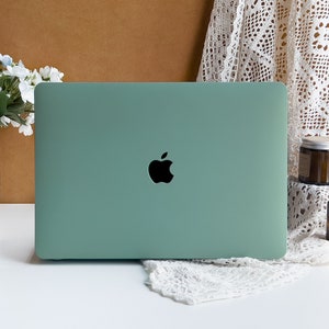 May include: A closed sage green laptop computer with an Apple logo on the lid. The laptop is sitting on a white surface with a white crocheted doily in front of it.