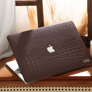 May include: A brown laptop case with a crocodile skin texture is placed on a wooden surface. The case has the Apple logo and gold initials "GM." A white candle and a window are in the background.