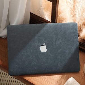 May include: A dark gray laptop case with the Apple logo in the center. The case has a textured appearance and is resting on a wooden surface. A white candle and a window are in the background.