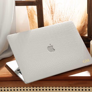 May include: A white laptop with a textured, crocodile-like pattern cover. The Apple logo is visible on the top. Gold initials "GM" are on the bottom right corner. The laptop is open on a wooden surface.