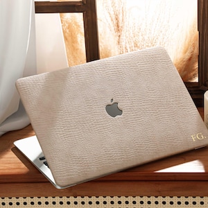 May include: A light beige laptop case with a textured surface, featuring the Apple logo and initials "FG." The laptop is open on a wooden surface, with a candle and window in the background. The case appears to be made of leather.