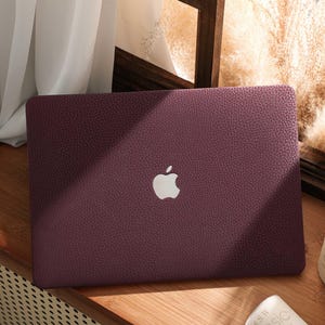 Puede incluir: Una funda de portátil burdeos con una superficie texturizada y un logotipo de Apple blanco. La funda está sobre una superficie de madera cerca de una ventana con una cortina blanca y una vela encendida.