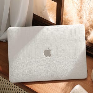 May include: A white laptop case with a textured, crocodile-skin pattern. The case has a cutout for the Apple logo. The laptop case is sitting on a wooden surface.
