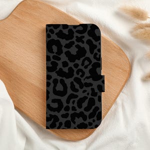May include: A black phone case with a dark gray leopard print pattern. The case is rectangular with a magnetic closure. The case is on a wooden cutting board with white fabric and dried plants in the background.