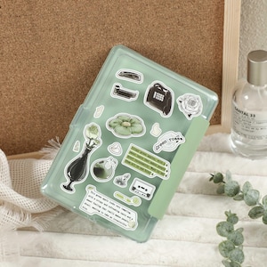 以下が含まれることがあります： ミントグリーンの透明なプラスチックケースに、植物やヴィンテージ風のデザインのステッカーが詰まっています。ステッカーには花瓶、カセットテープ、「green roses」の文字が含まれています。銀色のキャップが付いた透明なガラス瓶も写っています。