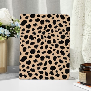 Puede incluir: Un cuaderno rectangular con un fondo beige y un estampado de leopardo negro. El cuaderno está de pie sobre una superficie blanca. Un pequeño tarro de vela marrón y una cesta de madera con flores están en el fondo.
