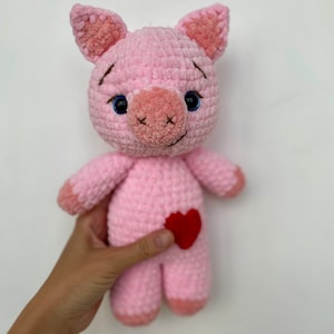Op de afbeelding: Een roze gehaakt knuffel varken met blauwe ogen en een rood vilt hartje op zijn buik.