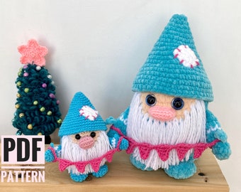 May include: Two crocheted gnomes with blue hats and white beards, holding a pink banner. The gnomes have large eyes and pink noses. A small green Christmas tree with colourful ornaments and a pink star topper is in the background. The text "PDF PATTERN" is in the lower left corner.