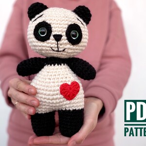 May include: Crochet amigurumi panda pattern PDF. The panda is white with black ears, eyes, and feet. It has a red heart on its chest.