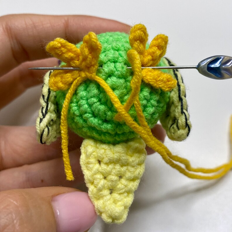 May include: Close-up of a crocheted green and yellow figure with yellow accents. A needle is inserted through the figure, with yellow yarn attached. The figure has a rounded head and a leaf-like shape at the bottom, suggesting a handmade craft project.