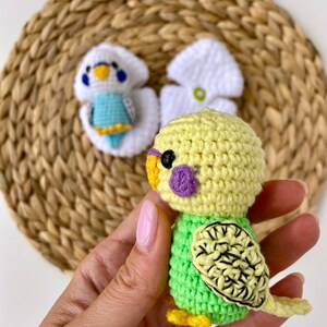 May include: A handmade crochet parakeet in shades of yellow and green, with a purple cheek patch and black eye. The bird has a detailed wing design and yellow feet. Two other crochet parakeets are in the background, one white and blue, and the other white.