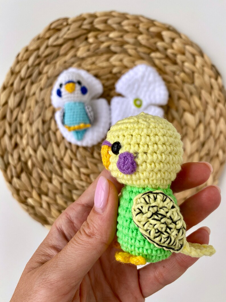 May include: A handmade crochet parakeet in shades of yellow and green, with a purple cheek patch and black eye. The bird has a detailed wing design and yellow feet. Two other crochet parakeets are in the background, one white and blue, and the other white.