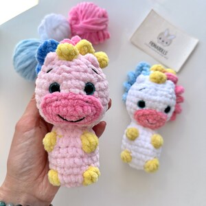 以下が含まれることがあります： ピンクと白の2つの編みぐるみユニコーンの玩具。黄色い毛糸の角と蹄があります。おもちゃは黒い目とピンクの鼻をしています。おもちゃは白い表面の上に置かれています。