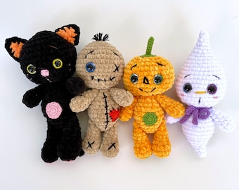 May include: Four crocheted Halloween toys: a black cat, a rag doll, a pumpkin, and a ghost. The cat has orange ears and pink accents. The rag doll has blue eyes and a red heart on its chest. The pumpkin has green accents and a smiling face. The ghost is white with purple accents and has blue eyes.
