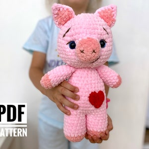 Op de afbeelding: Een roze gehaakt varkens knuffeldier met een rood hart op zijn borst. Het varken heeft blauwe ogen en een kleine roze snuit. Het knuffeldier wordt vastgehouden door een kinderhand.