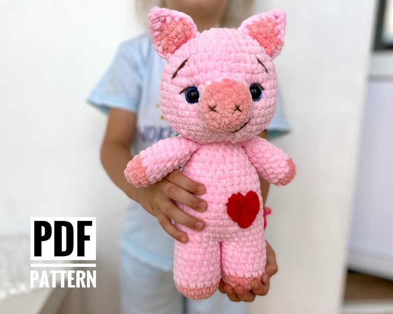 Op de afbeelding: Een roze gehaakt varkens knuffeldier met een rood hart op zijn borst. Het varken heeft blauwe ogen en een kleine roze snuit. Het knuffeldier wordt vastgehouden door een kinderhand.