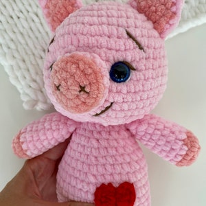 Op de afbeelding: Een roze gehaakt knuffelvarken met een rood hartje op zijn buik. Het varken heeft een blauw oog en een gestikte glimlach.