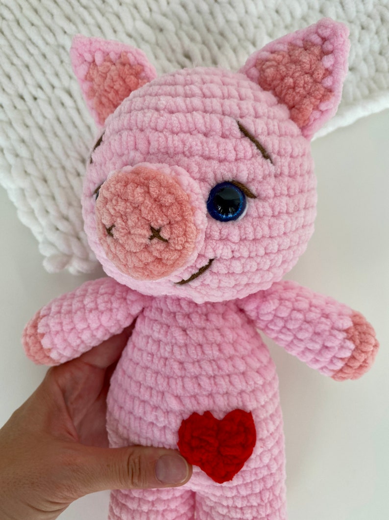 Op de afbeelding: Een roze gehaakt knuffelvarken met een rood hartje op zijn buik. Het varken heeft een blauw oog en een gestikte glimlach.