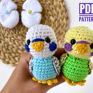 May include: Two crocheted parakeets, one blue and white, the other yellow and green, with black eyes and yellow beaks. The birds are handmade and have a detailed design. The image also includes two white crocheted egg-shaped items and text that reads "PDF PATTERN".