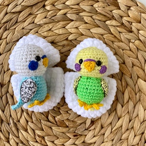 May include: Two handmade crochet parakeets. One is blue and white, the other is green and yellow. Both have white wings and are set against a woven, brown background. The parakeets have black eyes and yellow feet.