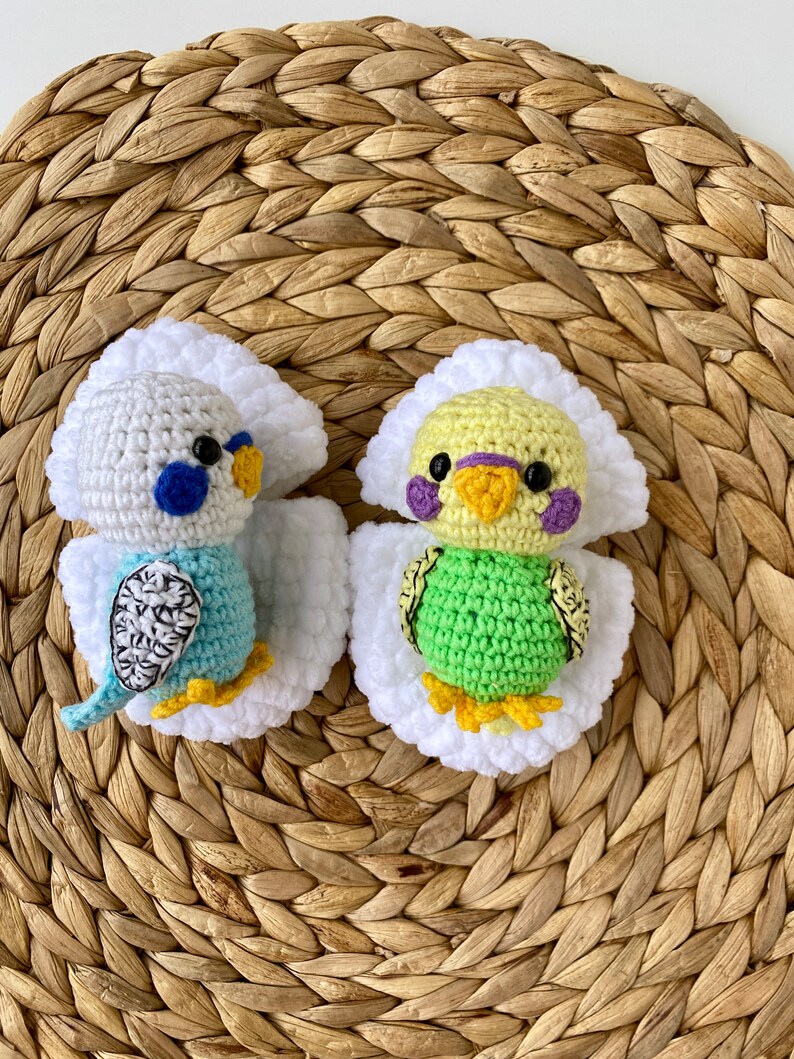 May include: Two handmade crochet parakeets. One is blue and white, the other is green and yellow. Both have white wings and are set against a woven, brown background. The parakeets have black eyes and yellow feet.