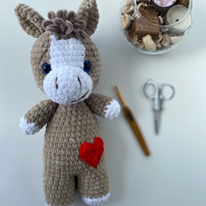 Op de afbeelding: Een gehaakte grijze en witte knuffel ezel met een rood hart op zijn borst. De ezel heeft blauwe ogen en een witte manen.