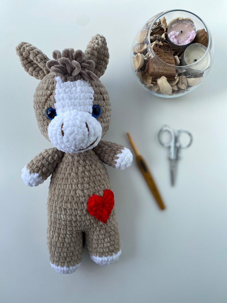 Op de afbeelding: Een gehaakte grijze en witte knuffel ezel met een rood hart op zijn borst. De ezel heeft blauwe ogen en een witte manen.