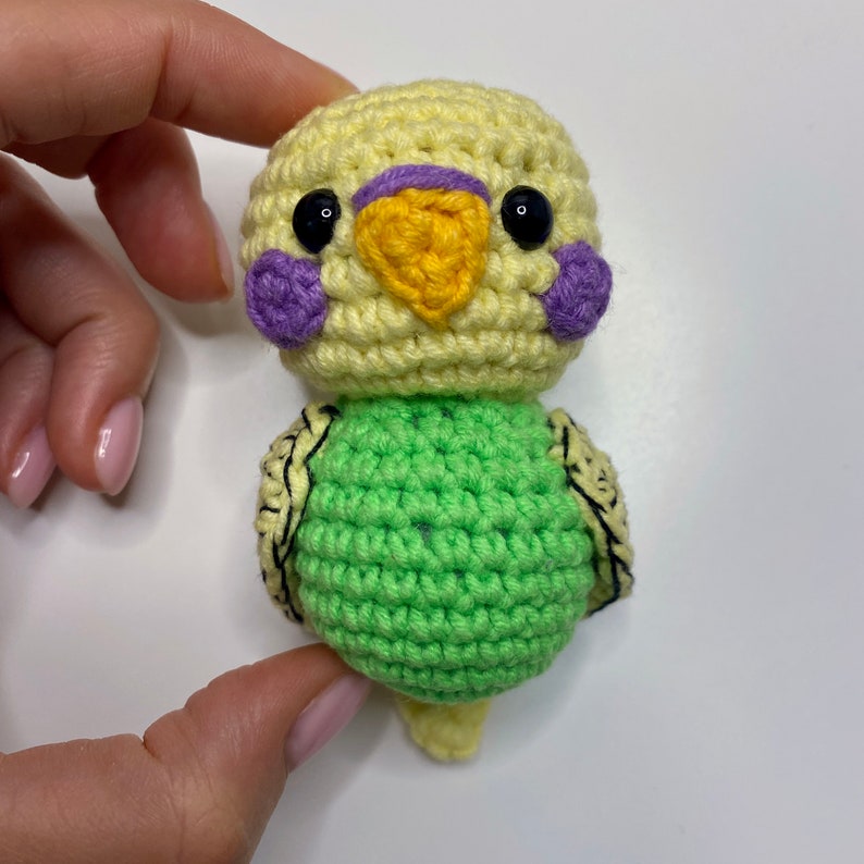 May include: A handmade crochet bird in yellow and green. The bird has a yellow head with purple cheek patches and a yellow beak. The body is green with black detailing. The bird is a small, soft toy.