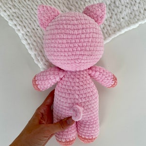 Op de afbeelding: Een roze gehaakt varkens knuffeldier met een gekrulde staart. Het varken staat op een wit oppervlak.