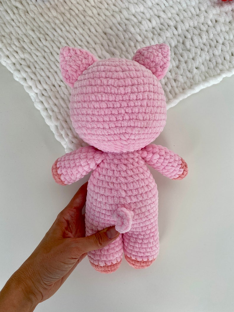Op de afbeelding: Een roze gehaakt varkens knuffeldier met een gekrulde staart. Het varken staat op een wit oppervlak.